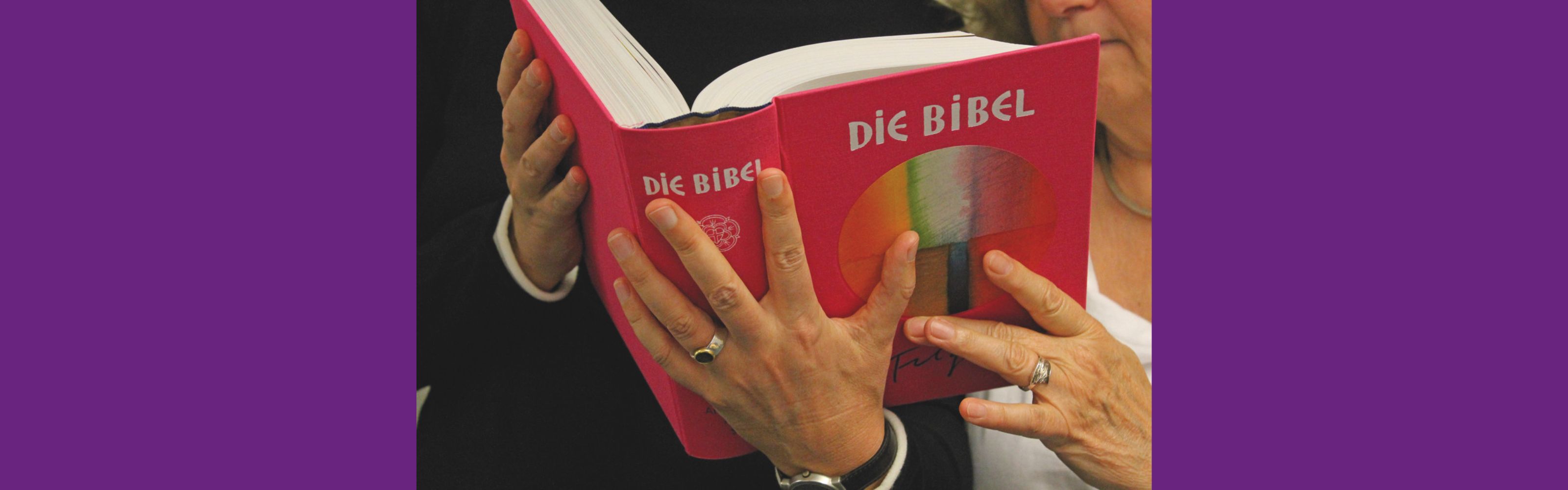 Zwei Frauen halten gemeinsam eine große, pinkfarbene Bibel mit der Aufschrift „DIE BIBEL“ in deutscher Sprache
