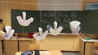 Gebastelte Friedenstauben hängen in einem Klassenzimmer vor einer Tafel mit Begriffen zum Pfingstfest