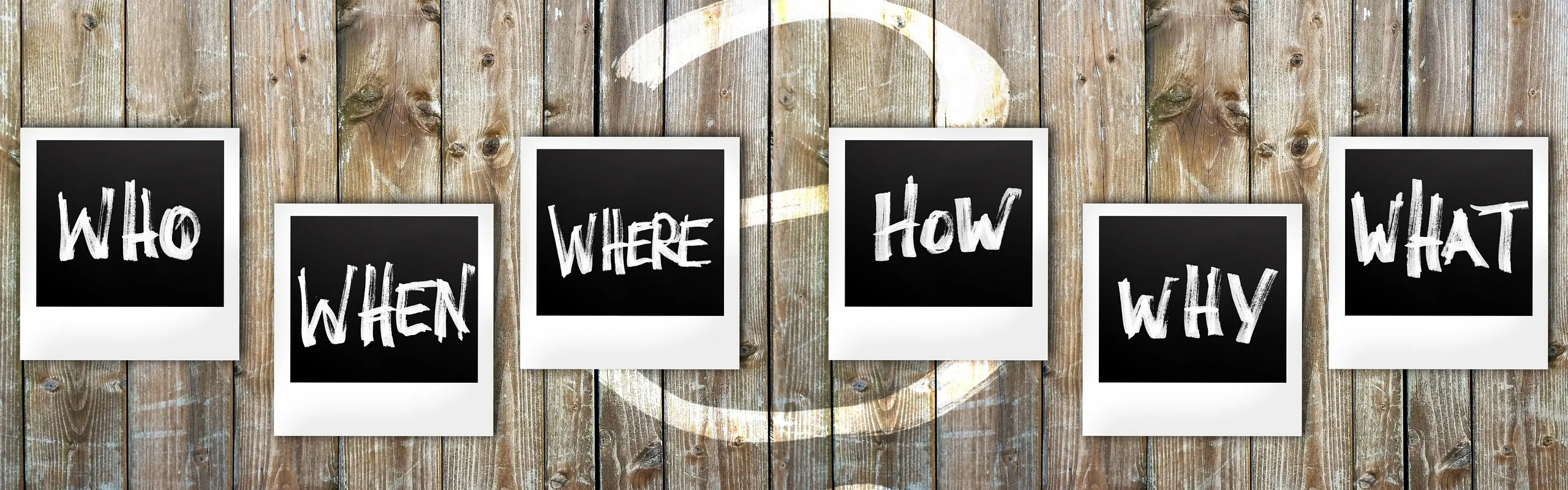 Sechs schwarze Tafeln mit den Fragen Who, When, Where, How, Why, What vor Holzwand