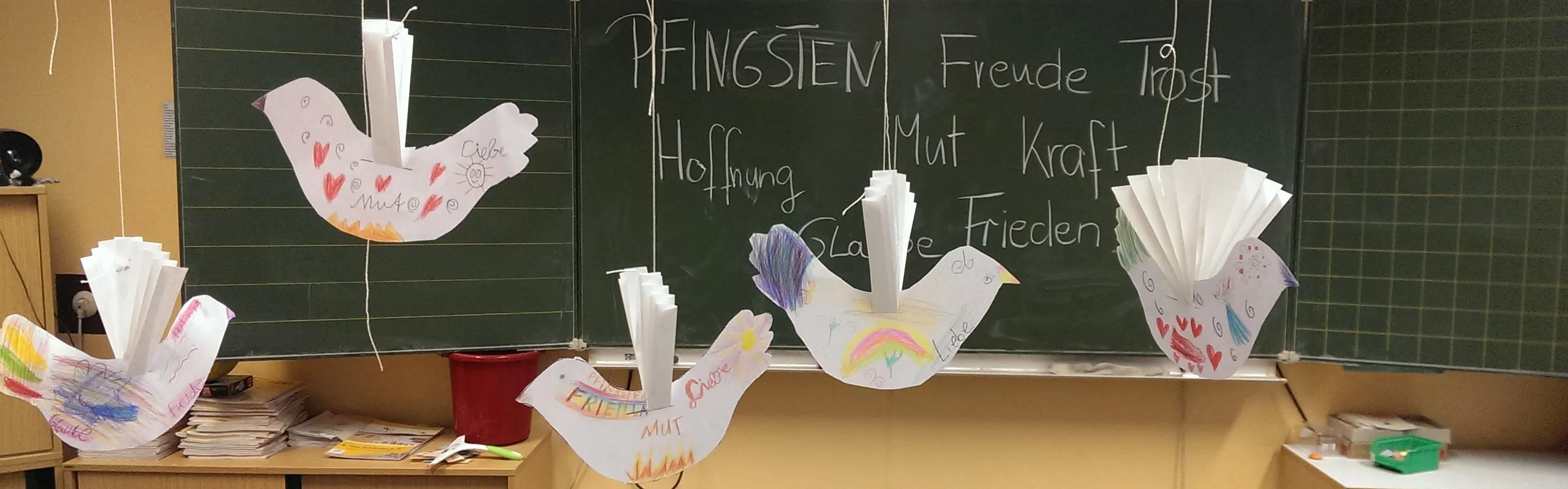 Gebastelte Friedenstauben hängen in einem Klassenzimmer vor einer Tafel mit Begriffen zum Pfingstfest