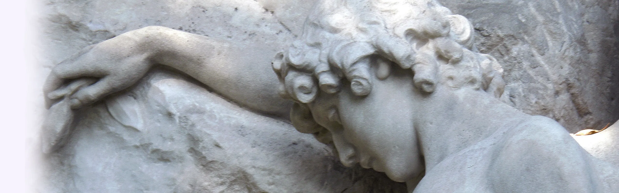 Steinfigur mit lockigem Haar beugt den Kopf traurig nach unten, stützt sich mit einem Arm an einem Felsen ab.