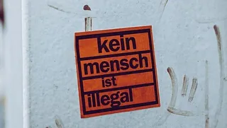 Orangefarbener Aufkleber an einer weißen Wand mit Text "kein mensch ist illegal"